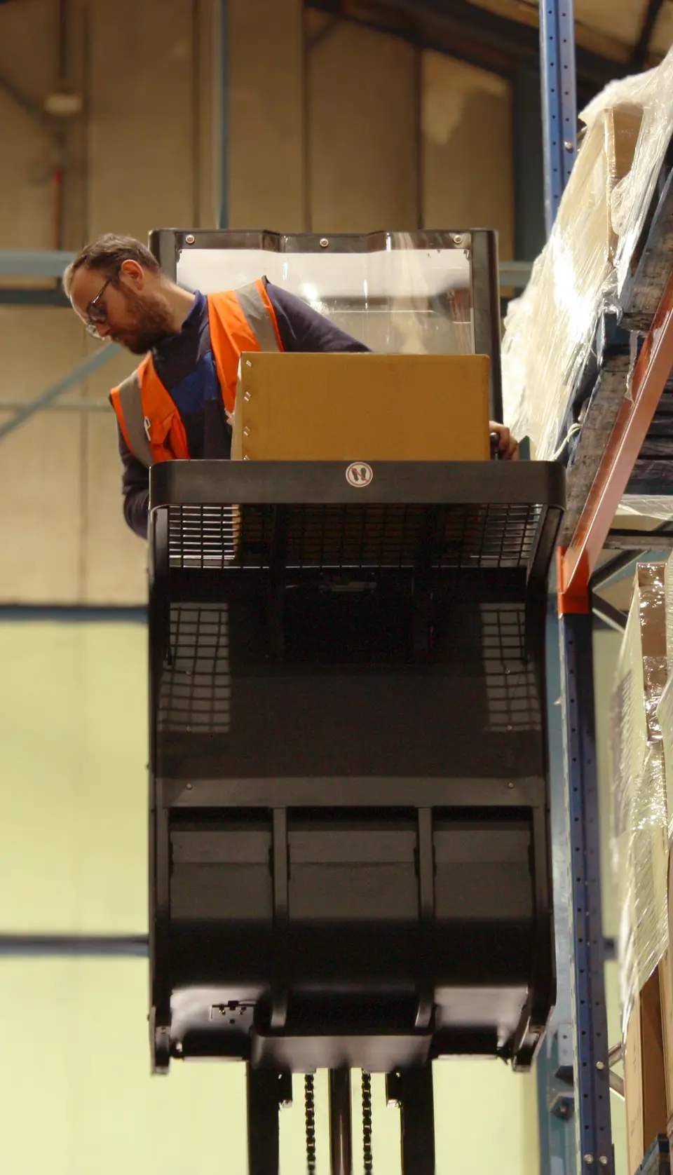Warehouse operative in high-visibility vest using an order picker forklift to retrieve a parcel from high racking shelves in a fulfilment centre.