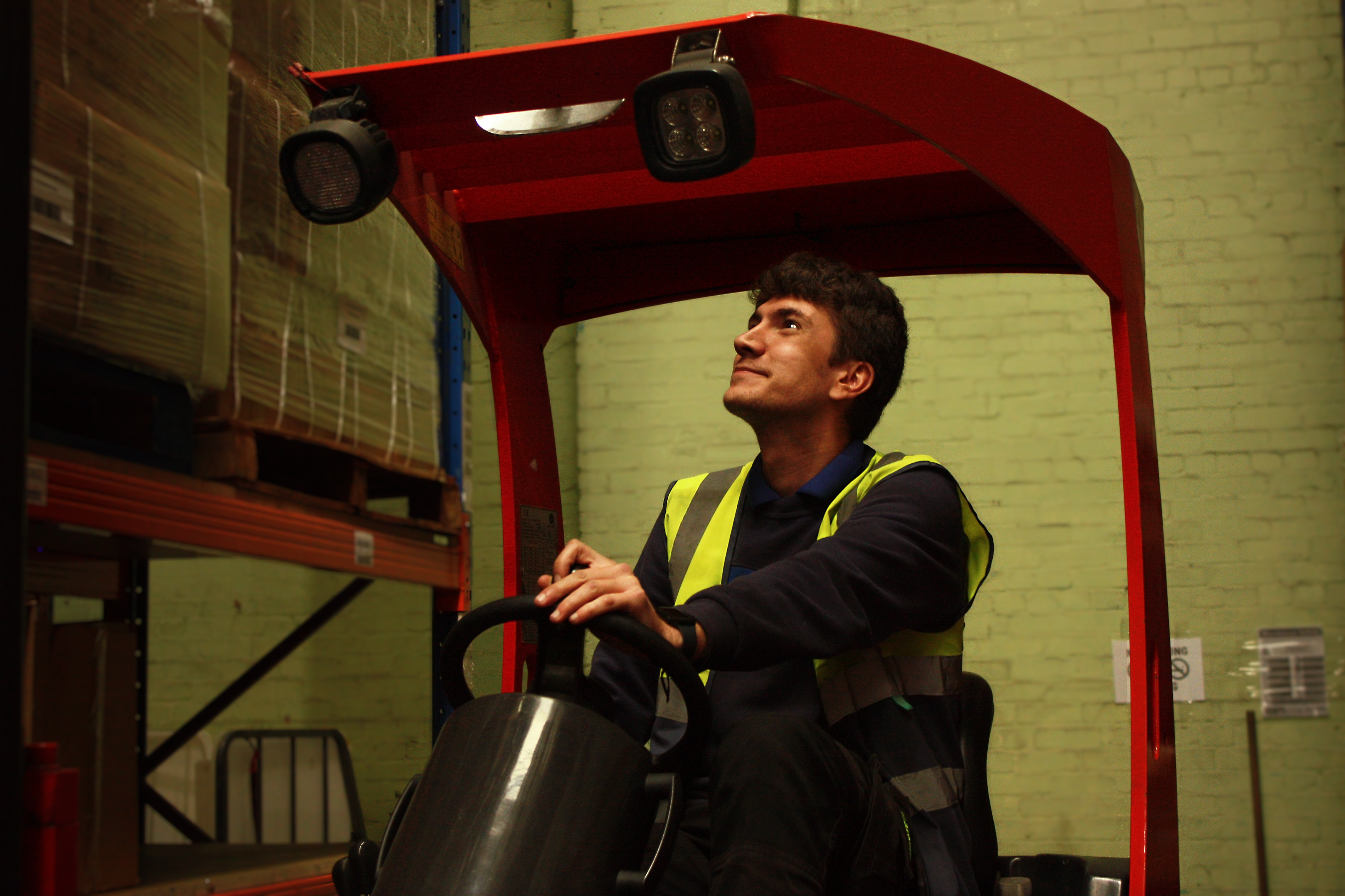 Forklift operator in a high-visibility vest looking up while manoeuvring stock in a fulfilment warehouse.